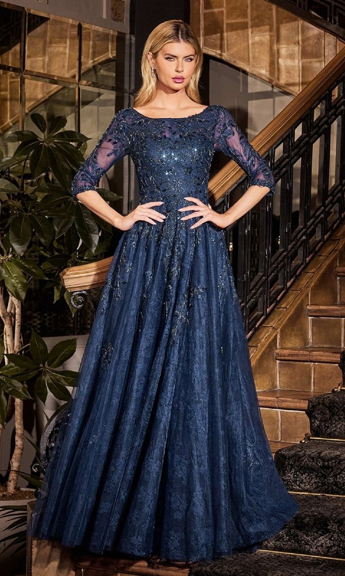 A woman stands on an indoor staircase wearing the Ladivine CK2401, a long sleeve blue lace formal gown with sheer sleeves and glitter details, ideal as a mother of the bride dress.